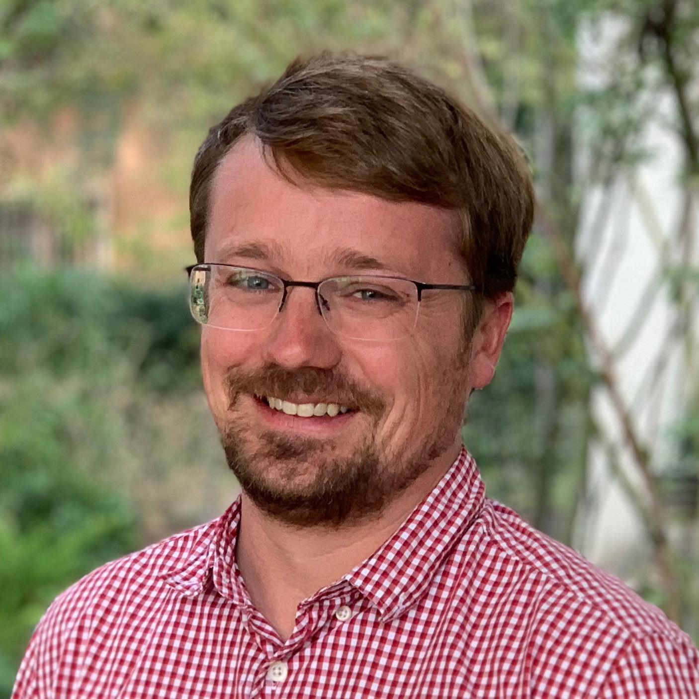 Austin's wearing glasses and a red and white checkered shirt in front of a blurred plant background. He's smiling.